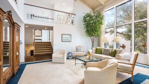 Amplia sala de estar con techo alto, una escalera que conduce a un nivel superior y grandes ventanales con vistas a árboles.