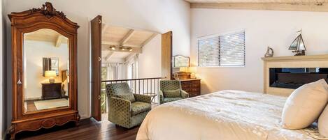A bedroom with a large mirror, a bed with white bedding, two armchairs, a dresser, and a fireplace.