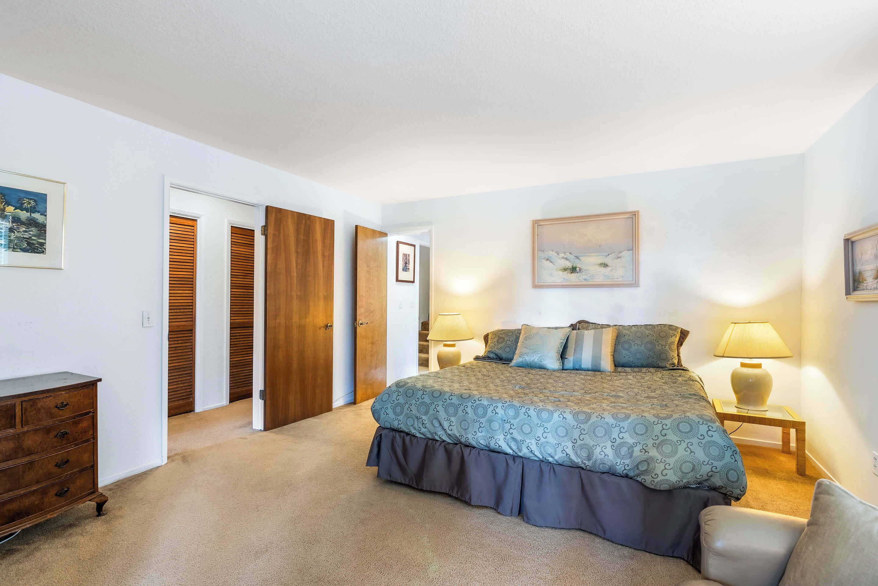 A bedroom with a large bed, a dresser, a nightstand, and a door leading to another room.