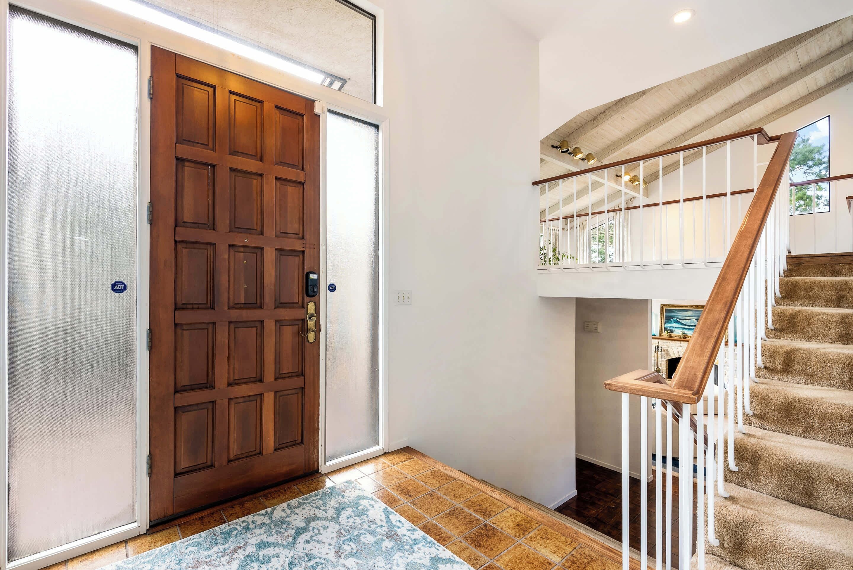 Une porte en bois avec un panneau de verre, un hall d’entrée recouvert d’un tapis et un escalier avec des rampes blanches menant à l’étage supérieur.