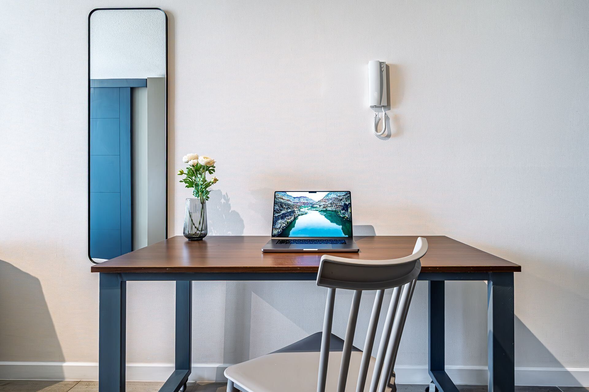 A modern room with a wooden desk, a laptop, a vase with flowers, and a wall-mounted telephone.