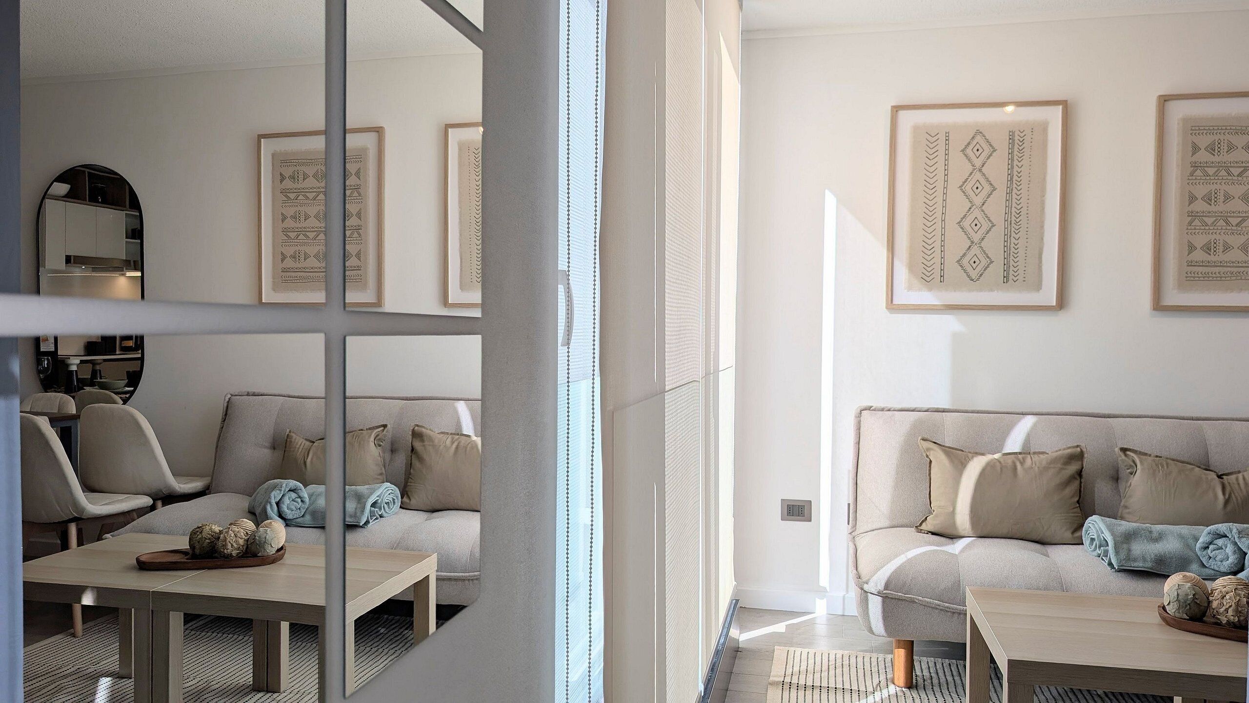Ein modernes Wohnzimmer mit Sofa, Sesseln und Couchtisch. An der Wand hängen gerahmte Kunstwerke, und durch eine Glaswand ist ein Küchenbereich erkennbar.