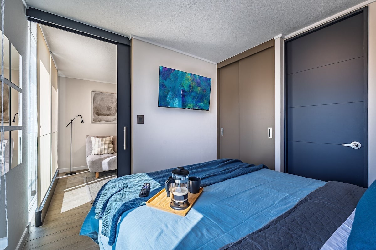 A modern bedroom with a bed, a coffee table with a coffee maker, a painting on the wall, and a sliding door.