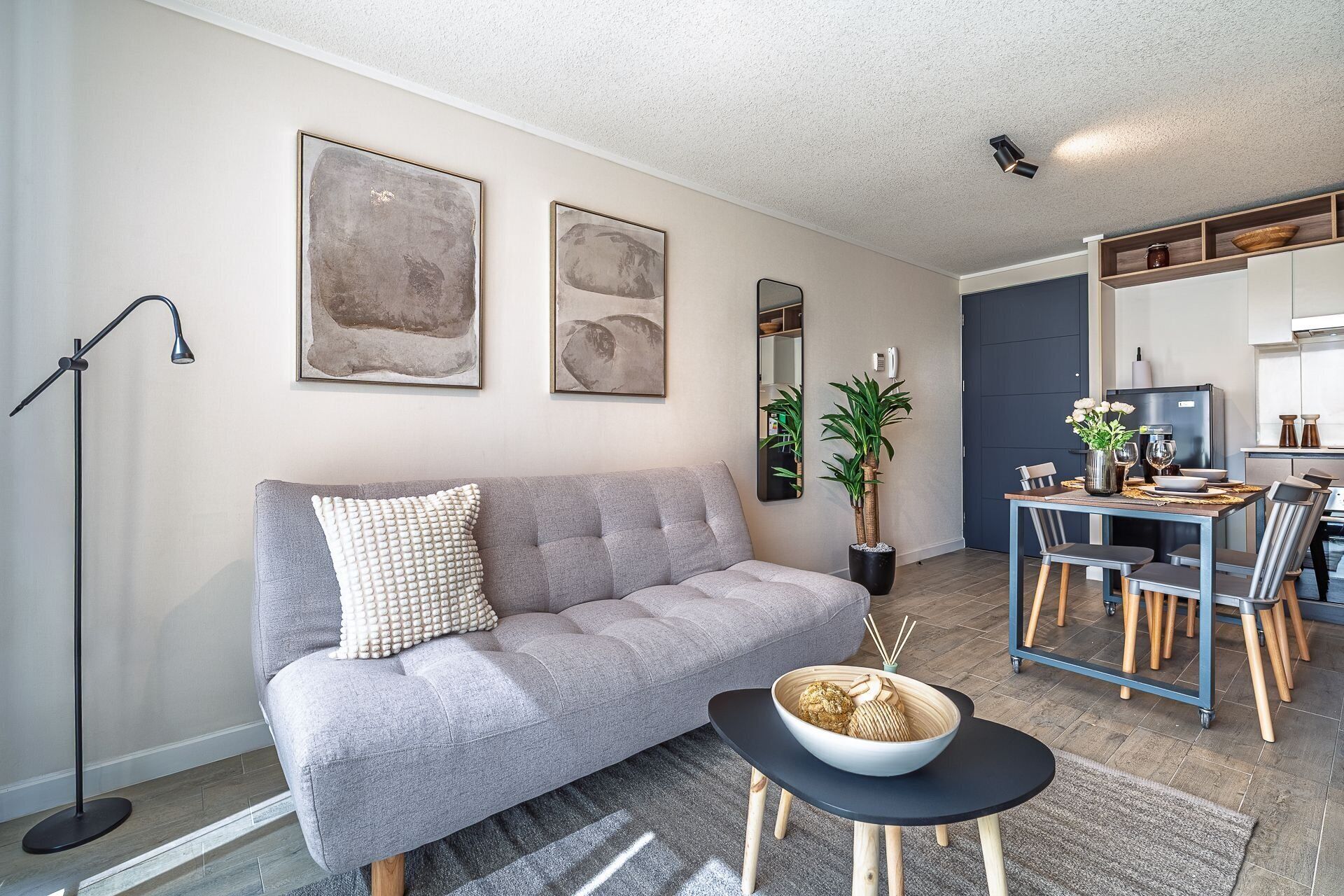 Una sala moderna con un sofá gris, una mesita redonda con un cuenco, una lámpara de pie y un comedor con una mesa de madera y sillas.