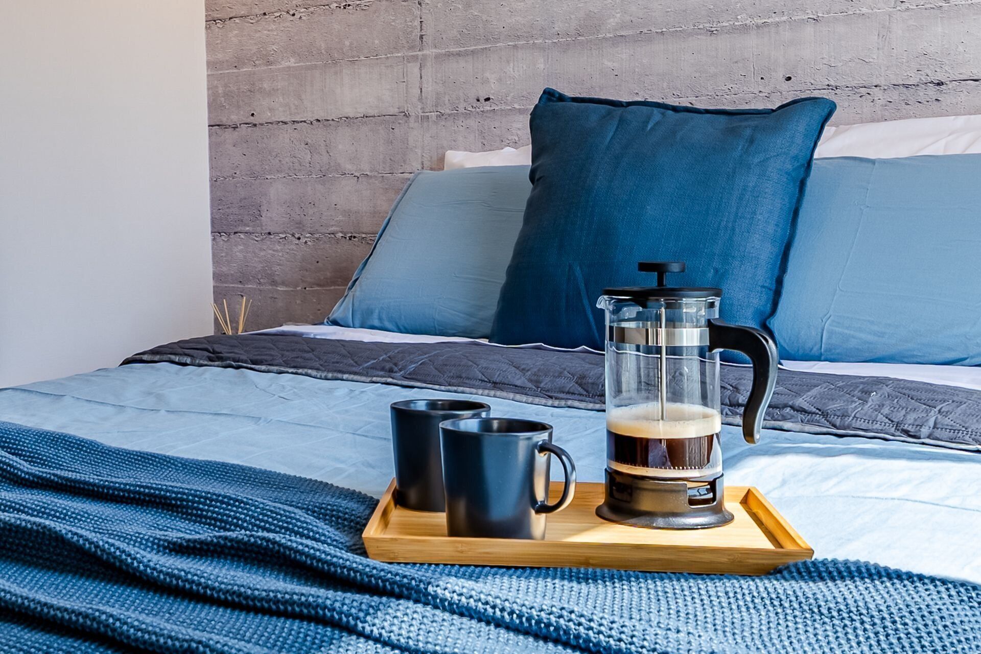 Un lit avec une couverture bleue, une machine à café et deux tasses noires sur un plateau.