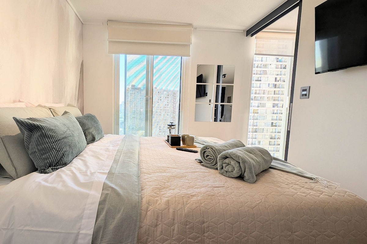A neatly made bed with pillows and a quilted blanket, a bedside table with a lamp, and a window with a view of buildings.