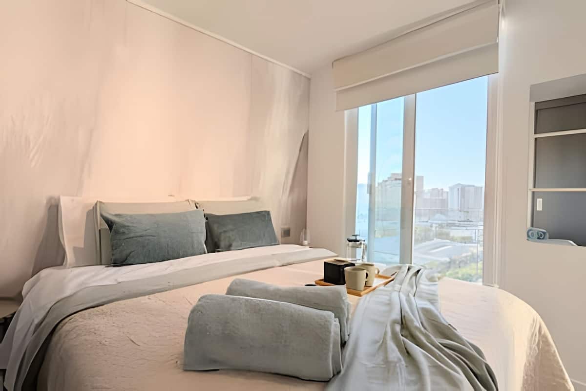 A modern hotel room with a large bed, a view of the city through floor-to-ceiling windows, and a small table with a cup and a book.