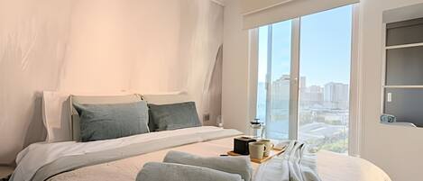 A modern hotel room with a large bed, a view of the city through floor-to-ceiling windows, and a small table with a cup and a book.