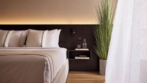 A modern bedroom with a large bed, a bedside table with a lamp, a small shelf with books, and a potted plant.