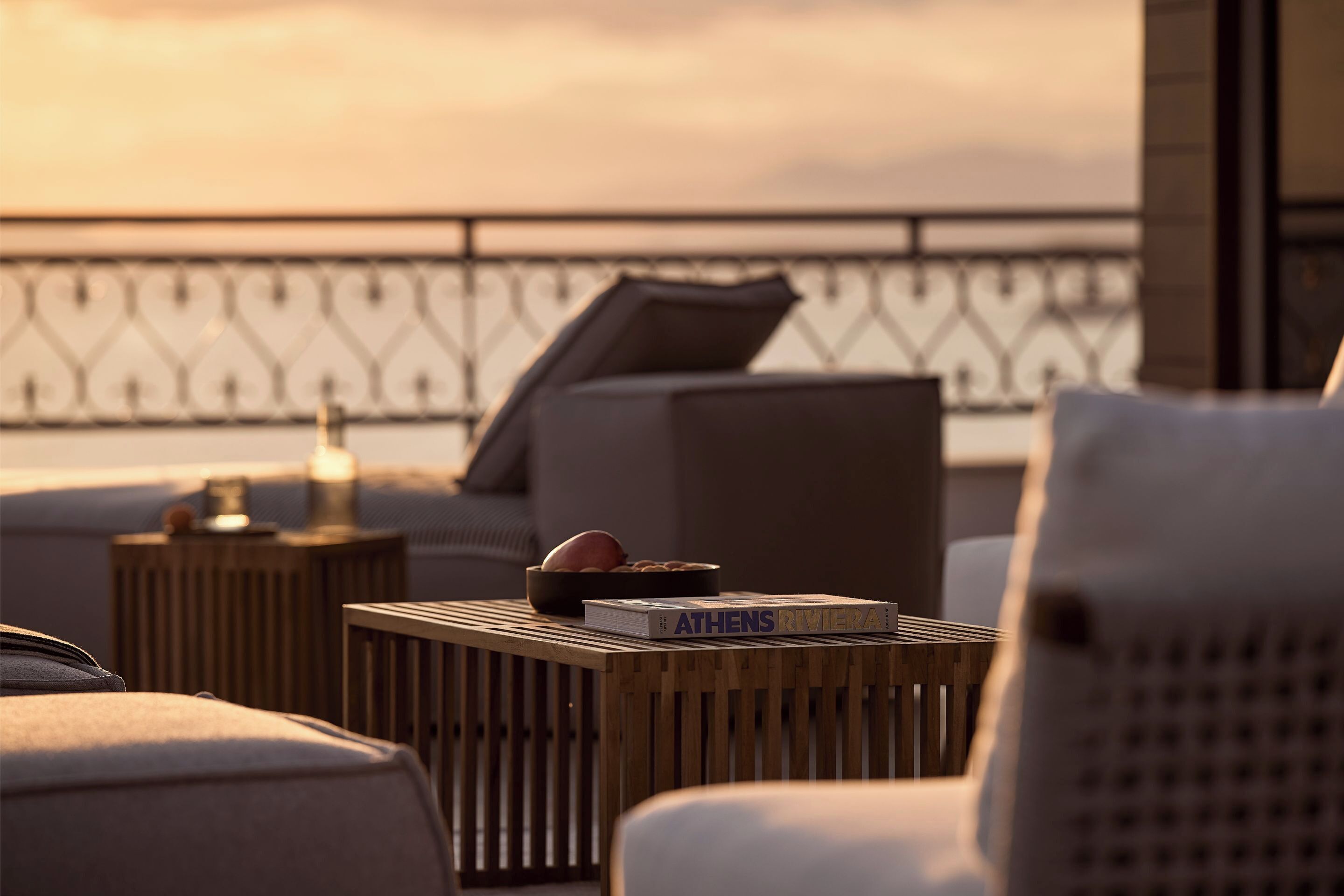 A modern outdoor seating area with a book titled "ATHENS RIVIERA" on a wooden table, a decorative bowl, and a lamp.