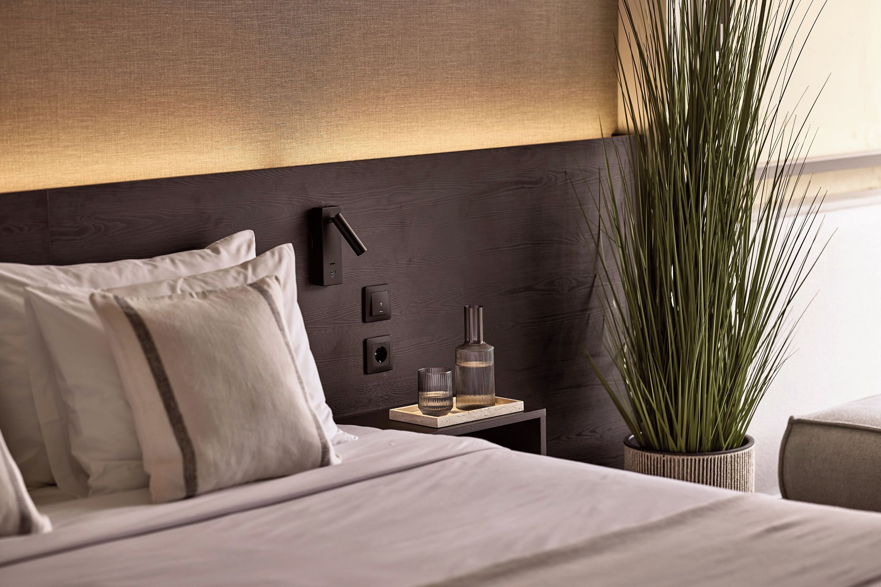 A neatly made bed with pillows, a bedside table with a lamp, a glass, and a bottle. There is a tall plant in a pot beside the bed.