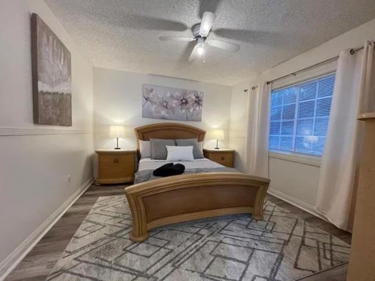 A bedroom with a bed, two bedside tables, a ceiling fan, a window with curtains, and a patterned rug.
