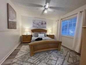 A bedroom with a bed, two bedside tables, a ceiling fan, a window with curtains, and a patterned rug.