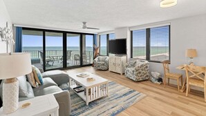 A spacious living room with a large sliding glass door leading to a balcony, a flat-screen TV, a sofa, a coffee table, and a dining area with a wooden table and chairs. - Book for Summer! Watercrest 709 sunset, huge balcony, beach service and towels! (Panama City Beach)