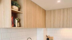 A modern kitchen with white cabinetry, a stainless steel dishwasher, a built-in oven, and a wooden shelf with decorative items. - Scandi City Escape | Walk Everywhere, Near Stadium (Christchurch)