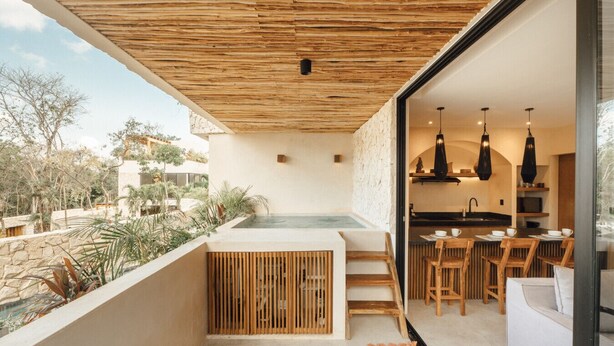 Una cucina esterna moderna con parete in pietra, posti a sedere in legno e vista su una piscina e vegetazione.