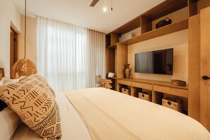A modern bedroom with a bed, a TV mounted on a wooden shelf, and a large window with sheer curtains.