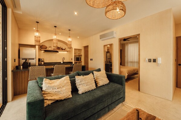A modern living room with a green sofa, wicker pendant lights, and a kitchen in the background.