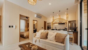 A modern living room with a sofa, a coffee table, and a kitchen area in the background. - 1BR Hidden Gem–Priv Pool+Garden at Launch Price (Tulum)