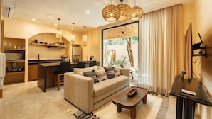 A modern living room with a sofa, coffee table, and a TV. There is a kitchen area with a dining table and chairs. The room has a large window with curtains. - Jungle Luxury: 1-BR, Plunge Pool, Fully Equipped (Tulum)