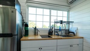 A kitchen with a stainless steel refrigerator, a green water dispenser, a sink with a knife block, and a window above the sink. - Container-Home 15 mins from FWTX | Pets Welcome (Fort Worth)