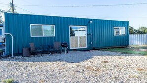 A blue mobile home with two windows and a sliding door. There are two chairs and a grill outside. - Container-Home 15 mins from FWTX | Pets Welcome (Fort Worth)