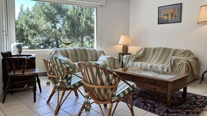 A living room with a sofa, two striped armchairs, a wooden coffee table, a lamp, and a view of trees through a window. - Large family house for 11 guests near the beach in Fréhel | Ref 113 (FREHEL)