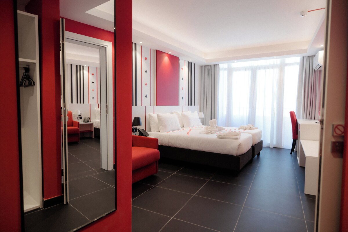 A modern hotel room with a large bed, a red armchair, a desk, and a large mirror.