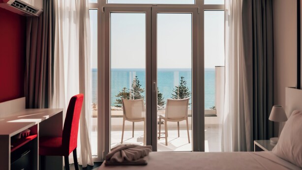 Une chambre d’hôtel avec un lit, un bureau, une chaise et une vue sur la mer, visible à travers de grandes portes vitrées.