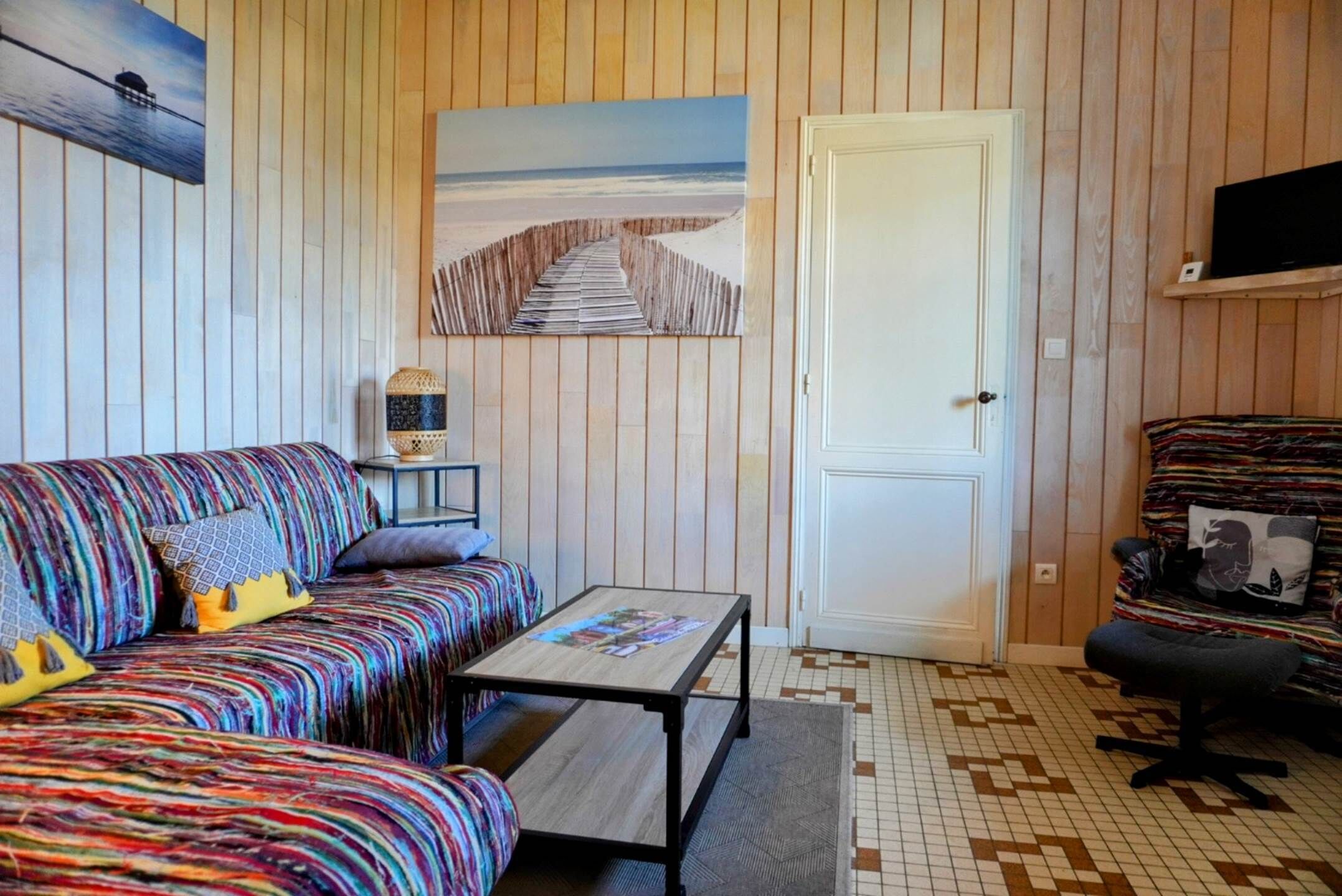 A room with a striped wall, a sofa with colorful cushions, a small table, a chair, and a door.