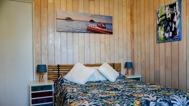 A bedroom with a wooden paneled wall, a bed with a floral bedspread, two bedside tables with lamps, and a painting of a boat on the wall.