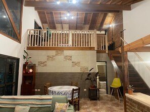 A cozy room with a wooden ceiling, a balcony, a dining area with a table and chairs, and a staircase.