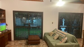 Un salon avec un canapé vert, un pouf marron, un bureau en bois avec un ordinateur et un téléviseur. De grandes fenêtres offrent une vue sur un jardin et une statue.