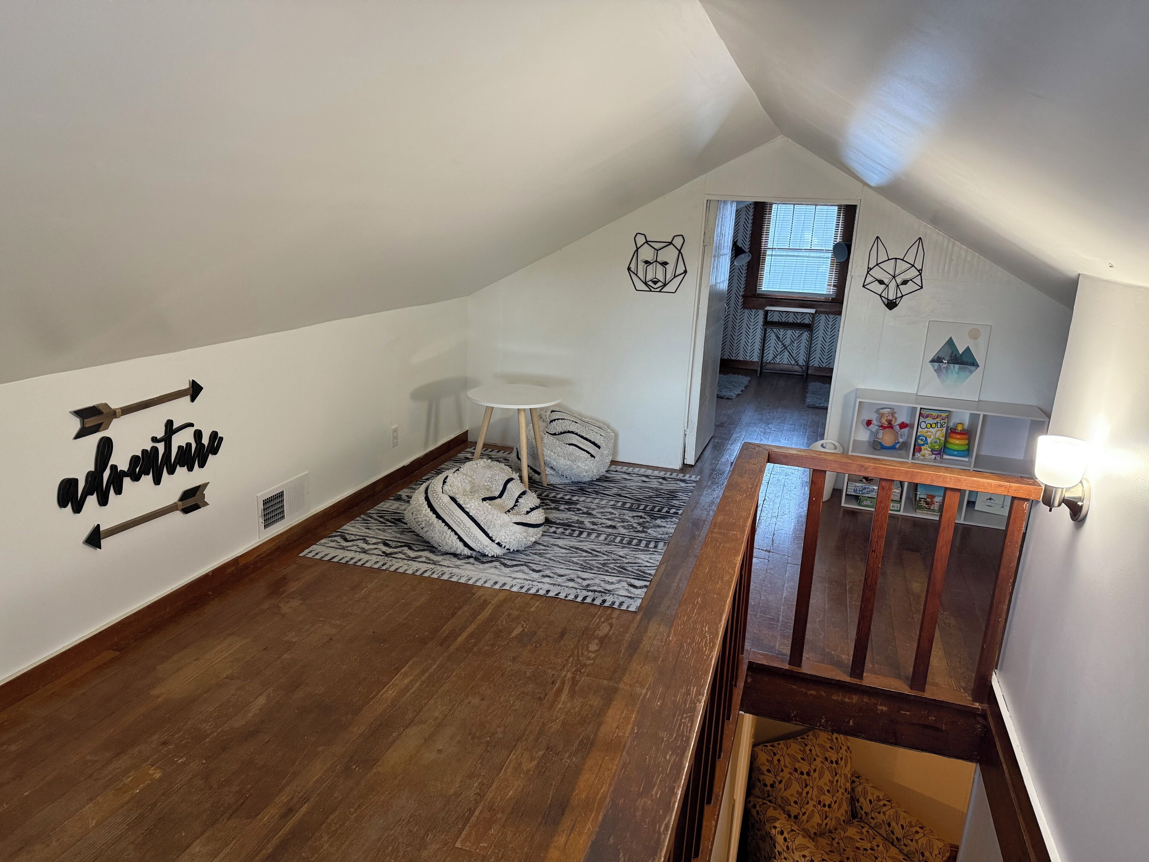 A cozy attic bedroom with a sloped ceiling, a wooden staircase, a white wall with a bear and cat design, a small round table, and a bookshelf with children's toys.