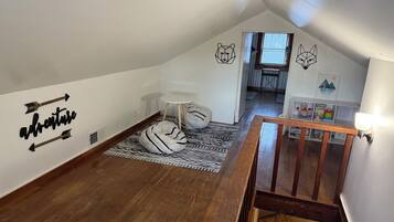 A cozy attic bedroom with a sloped ceiling, a wooden staircase, a white wall with a bear and cat design, a small round table, and a bookshelf with children's toys.
