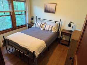 A neatly made bed with a white and gray blanket, a wooden headboard, and a bedside table with a lamp. There is a window with blinds, a framed picture on the wall, and a wooden dresser.