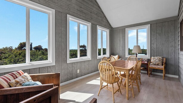 A dining area with a wooden table and chairs, large windows with a view of greenery, and a ceiling fan.