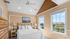 A bedroom with a bed, two bedside tables, a ceiling fan, a window with a view, and a painting on the wall.