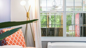Una habitación con un ventanal grande, un banco blanco, cojines de estampado naranja y una lámpara de pie.