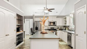 Una cocina moderna con armarios blancos, una isla central, un ventilador de techo y electrodomésticos de acero inoxidable.