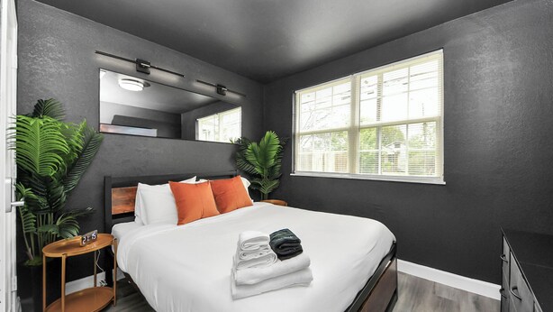 A modern bedroom with a large bed, a nightstand, a mirror, and a window with blinds.