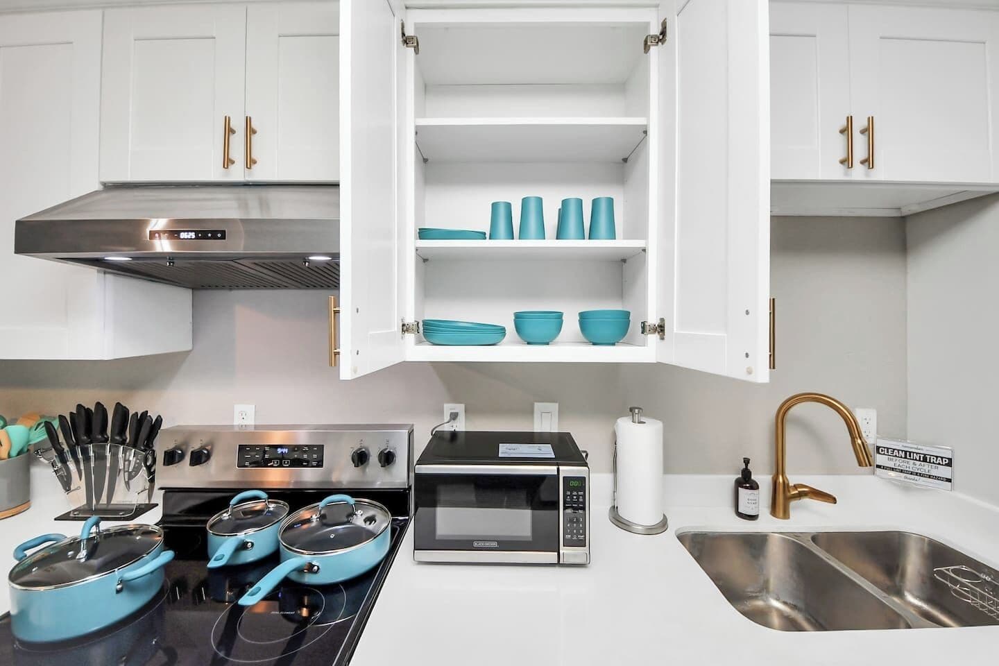 A modern kitchen with white cabinets, a stainless steel range hood, a microwave, a stove with pots, and a sink with a gold faucet.