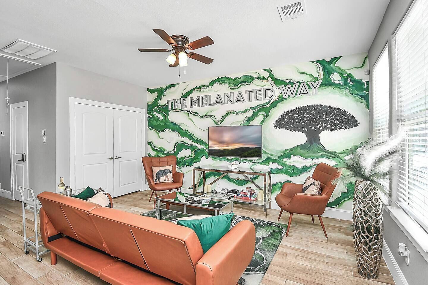 Una sala de estar con sofá, dos sillones, una mesa de centro, televisor y un mural con un árbol y texto.