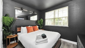 Quarto de hotel moderno com uma cama grande, uma mesa de cabeceira, um espelho, uma janela com persianas e plantas em vasos.