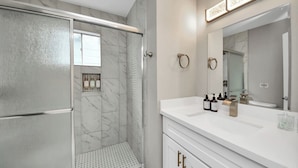 Casa de banho moderna com cabine de duche em vidro, toucador branco com um espelho grande e artigos de higiene no balcão.