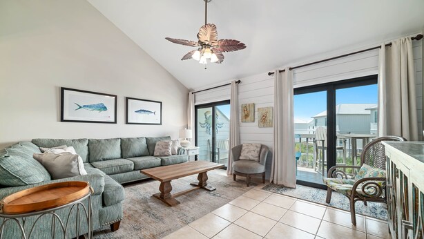 A spacious living room with a green sofa, a wooden coffee table, and a ceiling fan. There are framed pictures on the wall and a view of the outside through large windows.