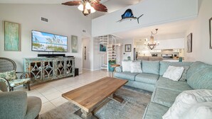 A spacious living room with a large wooden coffee table, a grey sofa, and a television. There is a ceiling fan, a staircase leading to an upper level, and a kitchen area visible in the background.