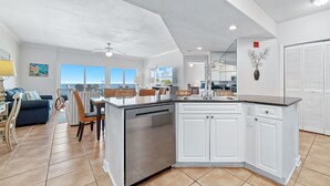 A modern kitchen with white cabinetry, a stainless steel dishwasher, and a dining area with wicker chairs. - Admiral Quarters 804 | Beachfront | Indoor & Outdoor Pools | Beach Getaways (Orange Beach)