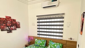 A bedroom with a bed, a ceiling fan, an air conditioner, a window with blinds, and a wall with a floral painting. - Magnificent T3 near the airport (Cotonou)