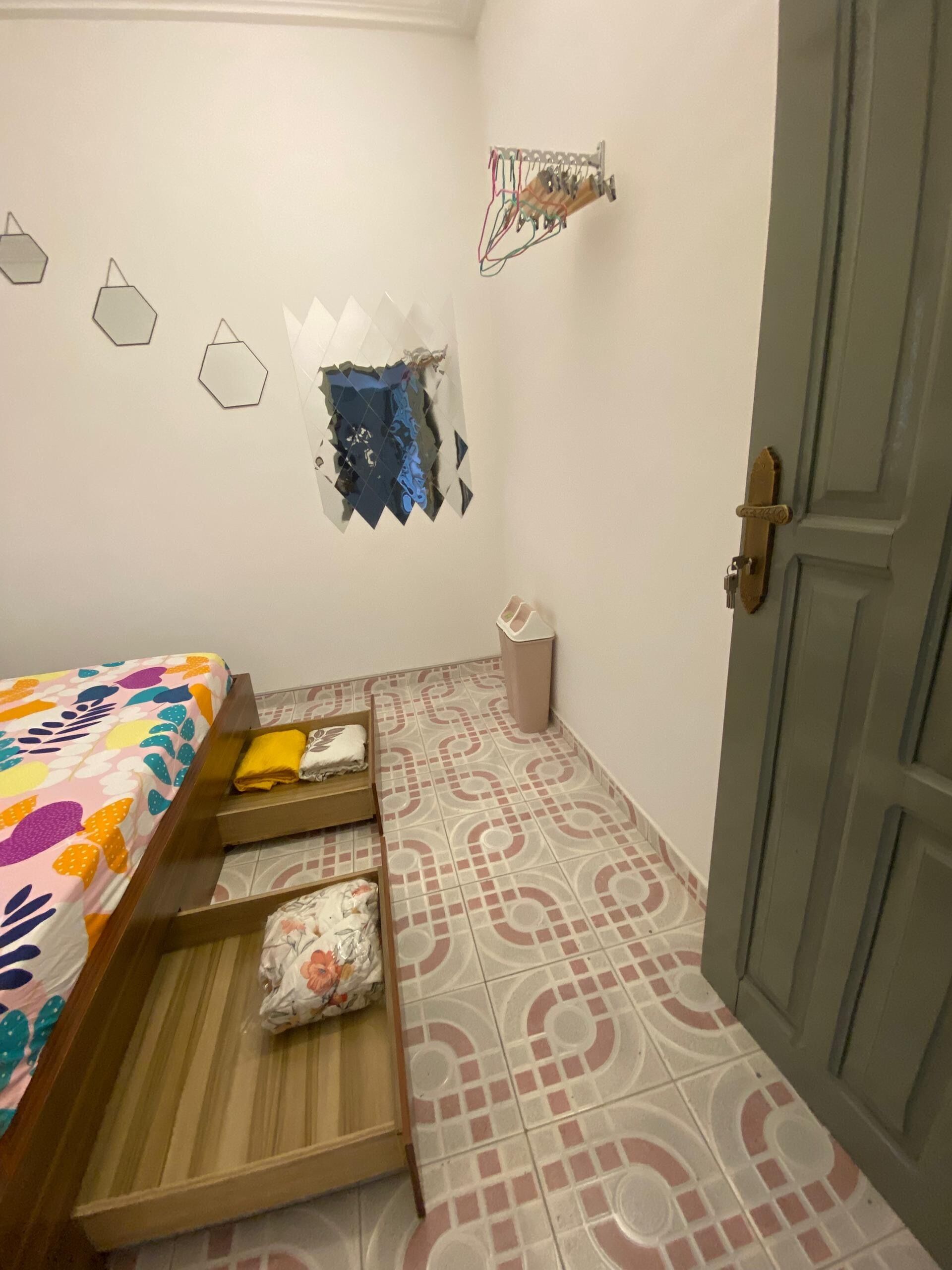 Een slaapkamer met een bed, een klein houten bankje, een prullenbak en een deur.